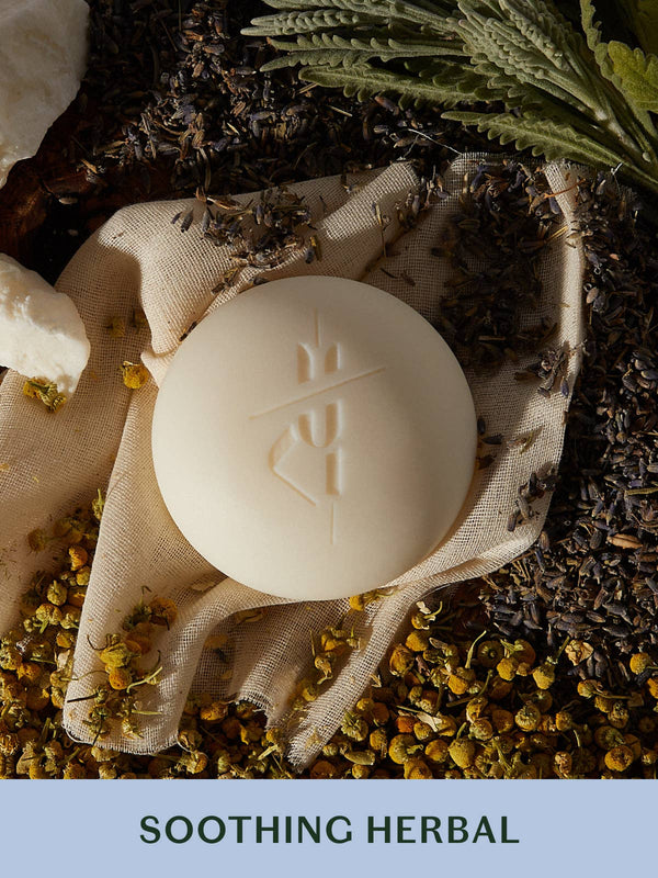 Bar of soap on a fabric with herbs and flowers, labeled 'Soothing Herbal'.