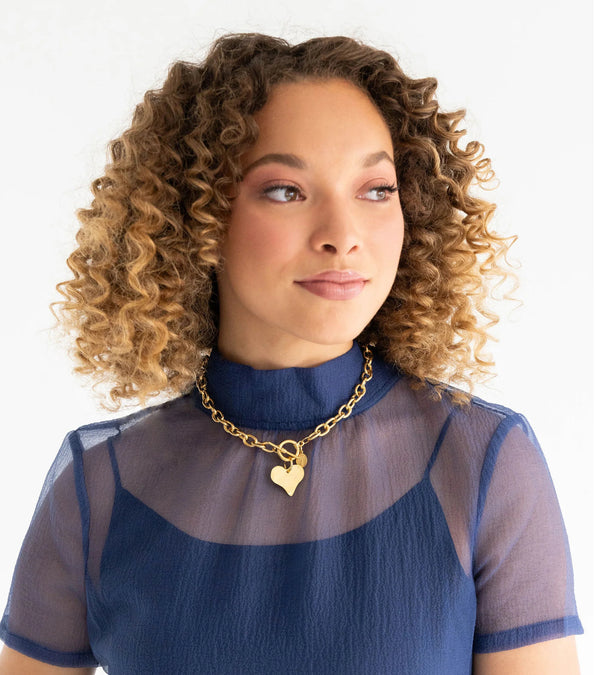 Woman wearing a blue top with a sheer overlay and a gold necklace with a heart pendant on a white background