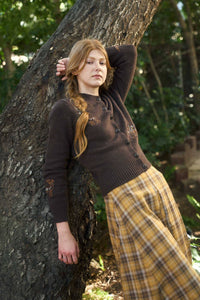 Woman in a brown sweater and yellow plaid skirt leaning against a tree in a forest setting